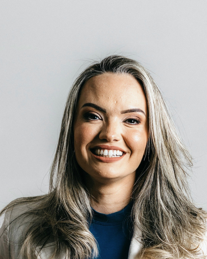 Retrato de estúdio de uma mulher sorrindo, de jaleco claro sobre blusa azul, em fundo cinza liso.