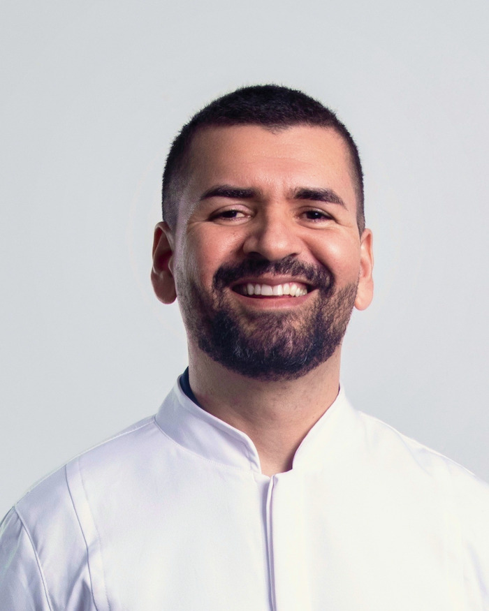 Retrato de estúdio de um homem sorrindo, de jaleco branco de gola alta, em fundo cinza claro liso.