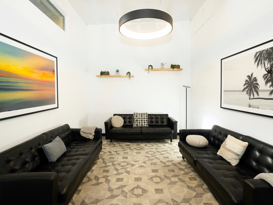 Sala de espera com dois sofás de couro preto, quadros grandes e luminária circular no teto.