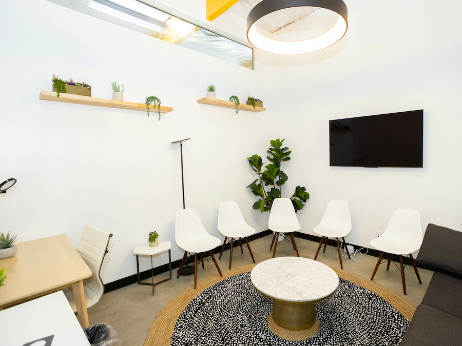Recepção clara, com mesa de madeira, cadeiras brancas em fila e plantas nas prateleiras.